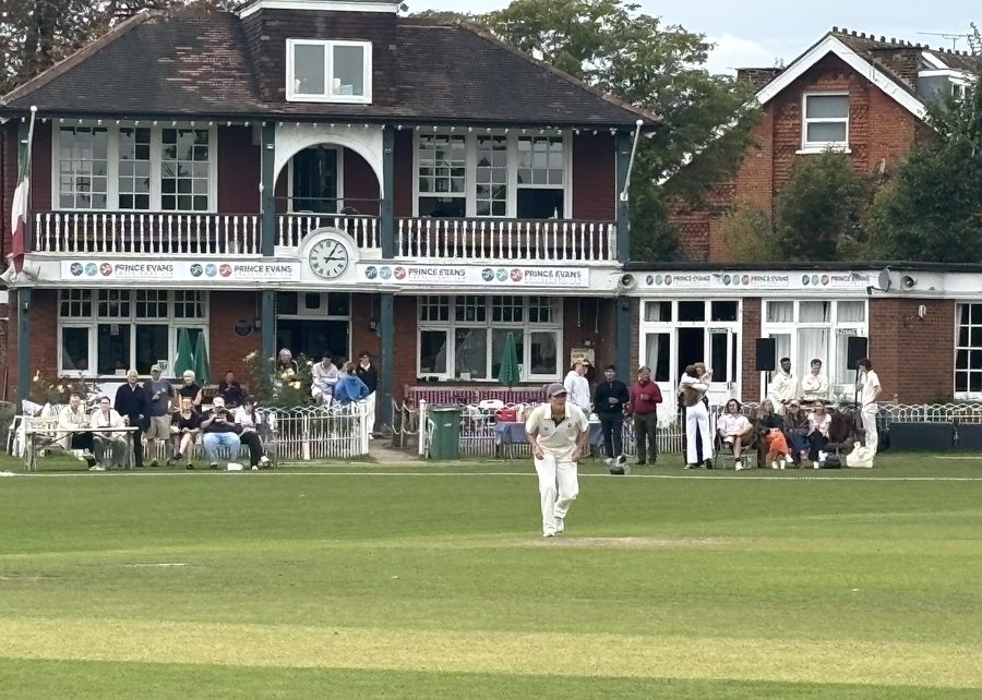Bowler running in to bowl, with spectators outside the pavilion